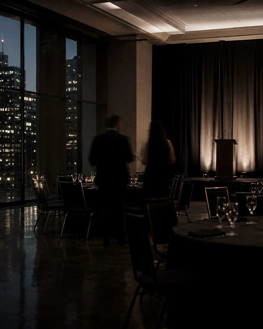 Ballroom set for a gala on a high floor, city towers lit beyond the window wall, an uplit lectern and screen at the far end, banquet rounds in the foreground